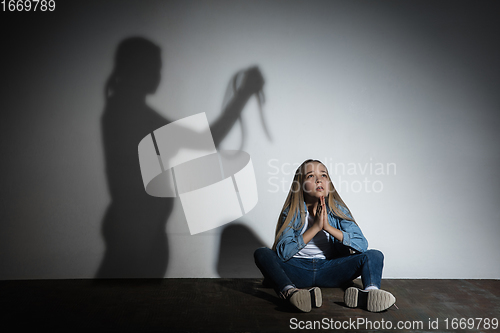 Image of Domestic violence, abusing. Scared little caucasian girl, victim sitting close to white wall with shadow of angry mother\'s threatening on it.