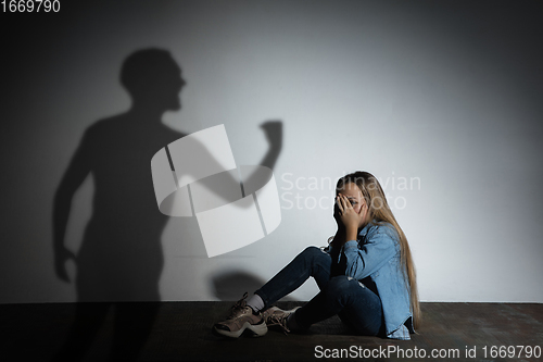 Image of Domestic violence, abusing. Scared little caucasian girl, victim sitting close to white wall with shadow of angry father\'s threatening on it.