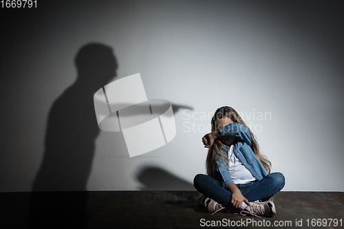 Image of Domestic violence, abusing. Scared little caucasian girl, victim sitting close to white wall with shadow of angry father\'s threatening on it.