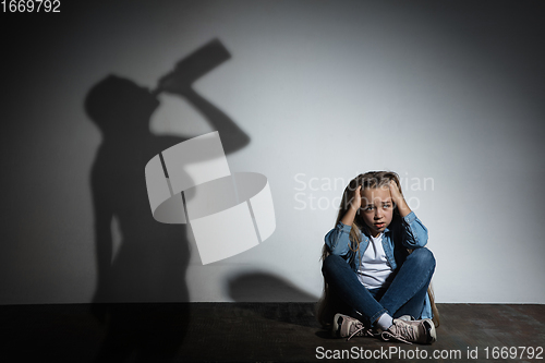 Image of Domestic violence, abusing. Scared little caucasian girl, victim sitting close to white wall with shadow of angry threatening mother with alcohol addiction.