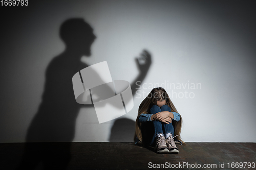 Image of Domestic violence, abusing. Scared little caucasian girl, victim sitting close to white wall with shadow of angry father\'s threatening on it.