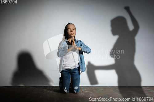 Image of Domestic violence, abusing. Scared little caucasian girl, victim sitting close to white wall with shadow of angry threatening mother with alcohol addiction.