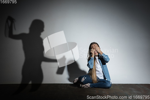 Image of Domestic violence, abusing. Scared little caucasian girl, victim sitting close to white wall with shadow of angry threatening mother with alcohol addiction.