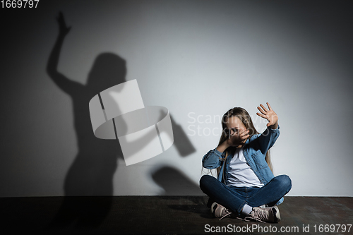 Image of Domestic violence, abusing. Scared little caucasian girl, victim sitting close to white wall with shadow of angry threatening mother with alcohol addiction.