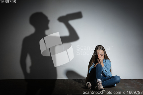 Image of Domestic violence, abusing. Scared little caucasian girl, victim sitting close to white wall with shadow of angry threatening father with alcohol addiction.