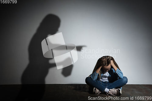 Image of Domestic violence, abusing. Scared little caucasian girl, victim sitting close to white wall with shadow of angry threatening mother with alcohol addiction.