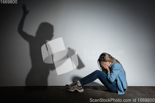 Image of Domestic violence, abusing. Scared little caucasian girl, victim sitting close to white wall with shadow of angry threatening mother with alcohol addiction.