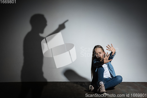 Image of Domestic violence, abusing. Scared little caucasian girl, victim sitting close to white wall with shadow of angry father\'s threatening on it.