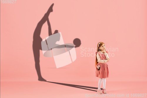 Image of Childhood and dream about big and famous future. Conceptual image with girl and shadow of fit female rhythmic gymnast on coral pink background