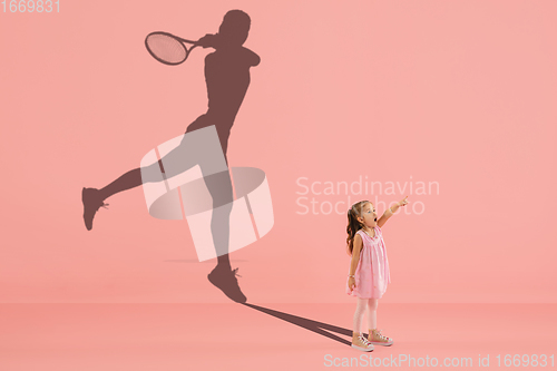 Image of Childhood and dream about big and famous future. Conceptual image with girl and shadow of fit female tennis player on coral pink background
