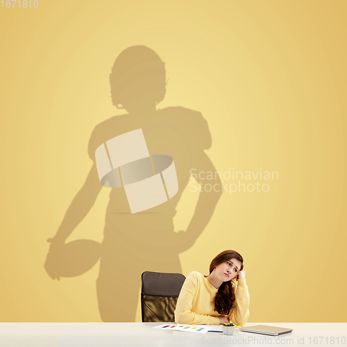 Image of Young woman dreaming about future in big sport during her work in office. Becoming a legend. Shadow of dreams on the wall behind her. Copyspace.