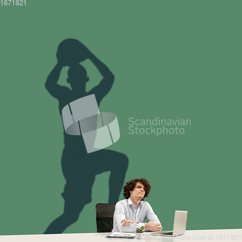 Image of Young man dreaming about future in big sport during his work in office. Becoming a legend. Shadow of dreams on the wall behind him. Copyspace.