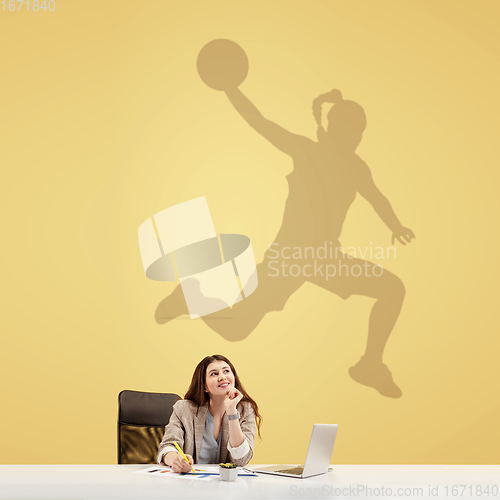 Image of Young woman dreaming about future in big sport during her work in office. Becoming a legend. Shadow of dreams on the wall behind her. Copyspace.