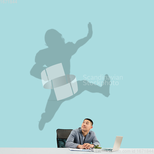 Image of Young man dreaming about future in big sport during his work in office. Becoming a legend. Shadow of dreams on the wall behind him. Copyspace.