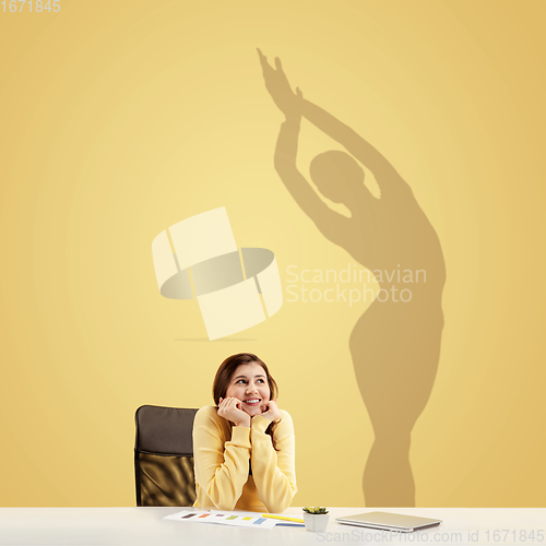 Image of Young woman dreaming about future in big sport during her work in office. Becoming a legend. Shadow of dreams on the wall behind her. Copyspace.