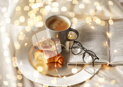 Image of croissants, cup of coffee, book and glasses in bed