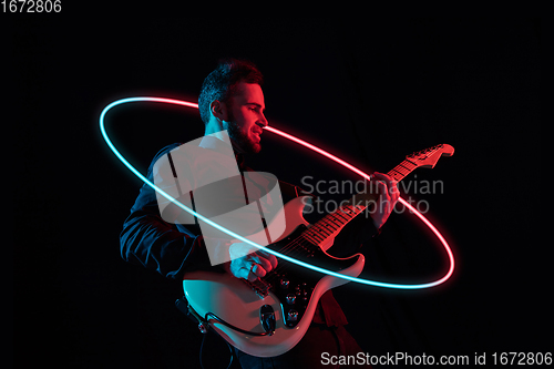 Image of Young and joyful caucasian musician playing guitar on black studio background in neon light with glowing neoned circle. Concept of music, hobby, festival.
