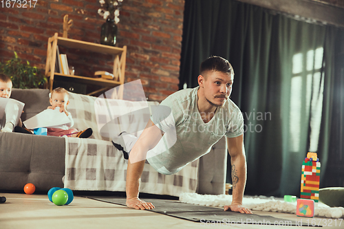 Image of Young man exercising fitness, aerobic, yoga at home, sporty lifestyle. Getting active while his child playing on the background, home gym.