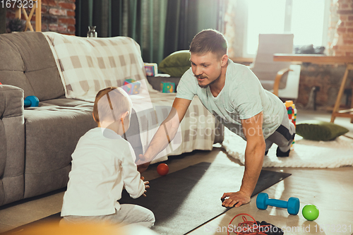 Image of Young man exercising fitness, aerobic, yoga at home, sporty lifestyle. Getting active while his child playing on the background, home gym.