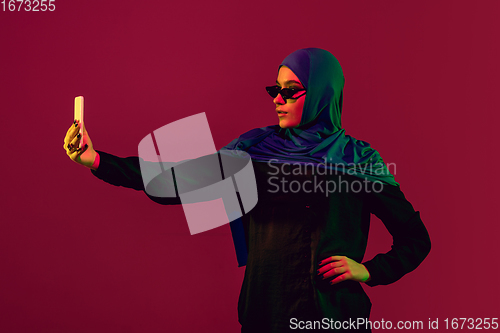 Image of Beautiful arab woman posing in stylish hijab isolated on burgundy studio background in neon light. Fashion, beauty, style concept