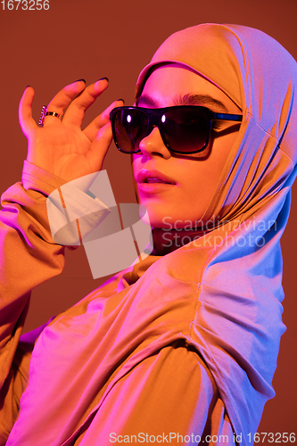 Image of Beautiful arab woman posing in stylish hijab isolated on brown studio background in neon light. Fashion, beauty, style concept