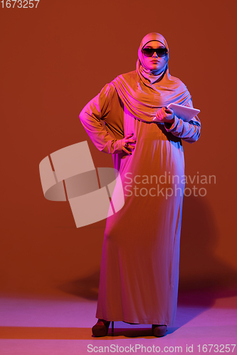 Image of Beautiful arab woman posing in stylish hijab isolated on brown studio background in neon light. Fashion, beauty, style concept