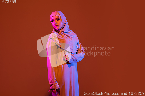 Image of Beautiful arab woman posing in stylish hijab isolated on brown studio background in neon light. Fashion, beauty, style concept