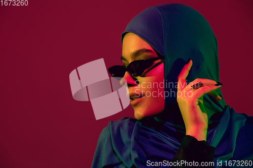 Image of Beautiful arab woman posing in stylish hijab isolated on burgundy studio background in neon light. Fashion, beauty, style concept