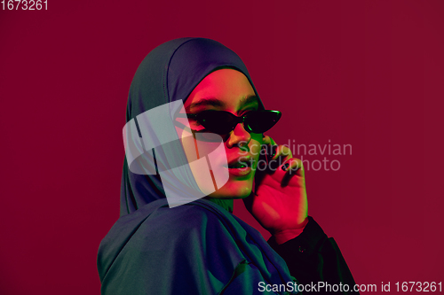 Image of Beautiful arab woman posing in stylish hijab isolated on burgundy studio background in neon light. Fashion, beauty, style concept