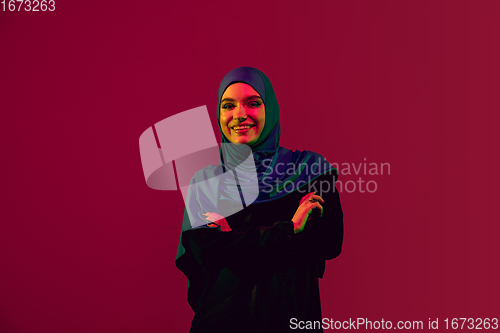 Image of Beautiful arab woman posing in stylish hijab isolated on burgundy studio background in neon light. Fashion, beauty, style concept