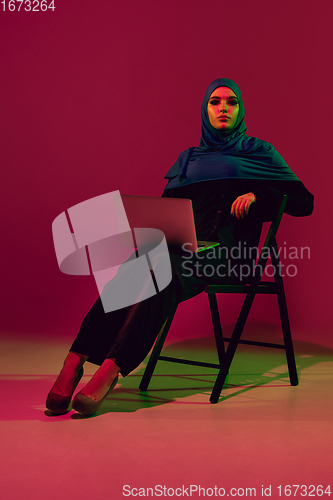 Image of Beautiful arab woman posing in stylish hijab isolated on burgundy studio background in neon light. Fashion, beauty, style concept