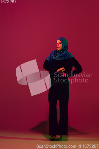 Image of Beautiful arab woman posing in stylish hijab isolated on burgundy studio background in neon light. Fashion, beauty, style concept