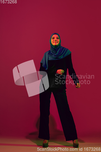 Image of Beautiful arab woman posing in stylish hijab isolated on burgundy studio background in neon light. Fashion, beauty, style concept