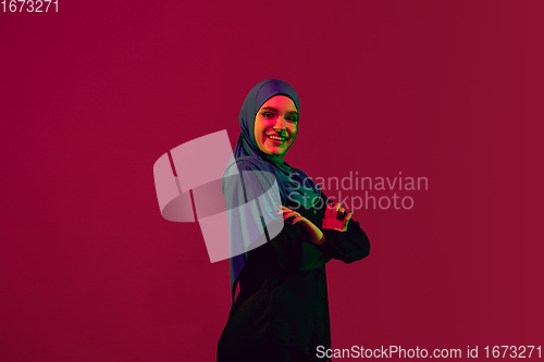 Image of Beautiful arab woman posing in stylish hijab isolated on burgundy studio background in neon light. Fashion, beauty, style concept