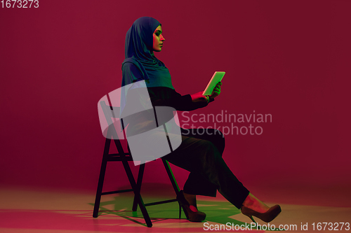 Image of Beautiful arab woman posing in stylish hijab isolated on burgundy studio background in neon light. Fashion, beauty, style concept