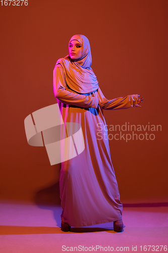 Image of Beautiful arab woman posing in stylish hijab isolated on brown studio background in neon light. Fashion, beauty, style concept