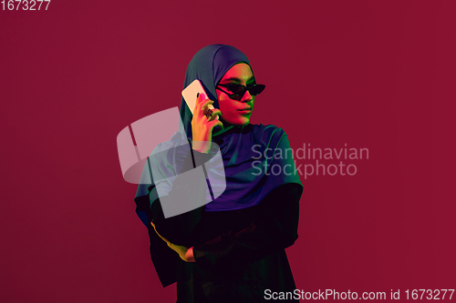 Image of Beautiful arab woman posing in stylish hijab isolated on burgundy studio background in neon light. Fashion, beauty, style concept