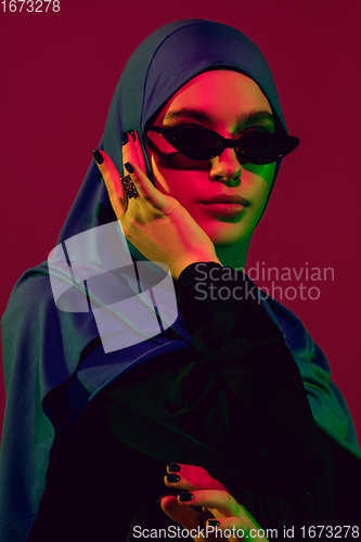 Image of Beautiful arab woman posing in stylish hijab isolated on burgundy studio background in neon light. Fashion, beauty, style concept