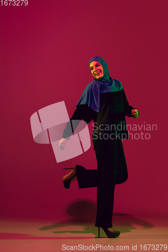 Image of Beautiful arab woman posing in stylish hijab isolated on burgundy studio background in neon light. Fashion, beauty, style concept