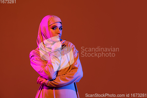 Image of Beautiful arab woman posing in stylish hijab isolated on brown studio background in neon light. Fashion, beauty, style concept