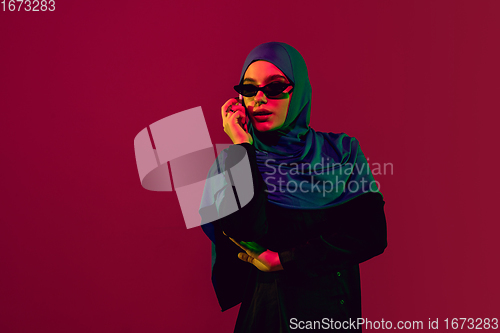 Image of Beautiful arab woman posing in stylish hijab isolated on burgundy studio background in neon light. Fashion, beauty, style concept