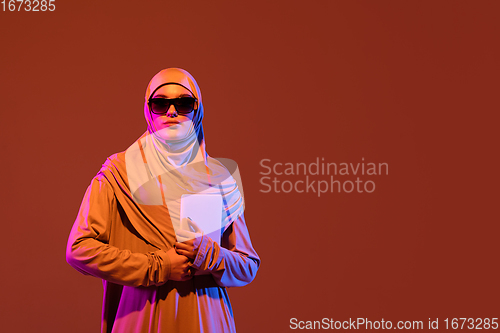 Image of Beautiful arab woman posing in stylish hijab isolated on brown studio background in neon light. Fashion, beauty, style concept