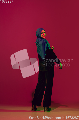 Image of Beautiful arab woman posing in stylish hijab isolated on burgundy studio background in neon light. Fashion, beauty, style concept