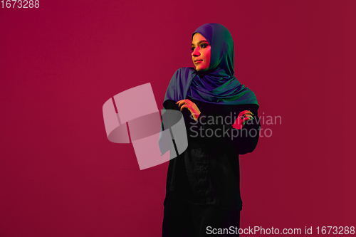 Image of Beautiful arab woman posing in stylish hijab isolated on burgundy studio background in neon light. Fashion, beauty, style concept