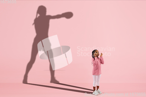 Image of Childhood and dream about big and famous future. Conceptual image with girl and shadow of fit female boxer on coral pink background