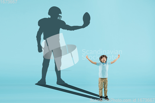 Image of Childhood and dream about big and famous future. Conceptual image with boy and shadow of fit male football player on blue background