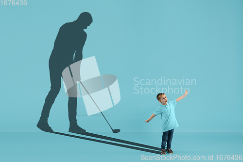 Image of Childhood and dream about big and famous future. Conceptual image with boy and shadow of male golf player on blue background