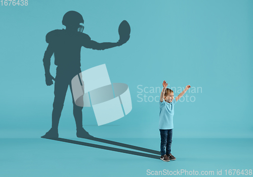 Image of Childhood and dream about big and famous future. Conceptual image with boy and shadow of american football player on blue background