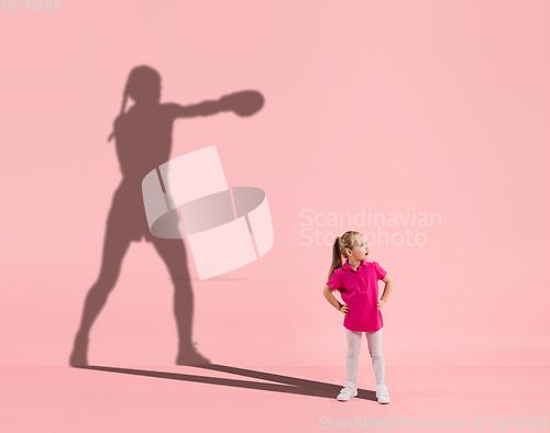 Image of Childhood and dream about big and famous future. Conceptual image with girl and shadow of fit female boxer on coral pink background