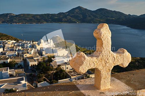 Image of Christian cross and Plaka village on Milos island over red geranium flowers on sunset in Greece